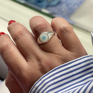 Op de afbeelding: Een zilveren ring met een oogontwerp, met een lichtblauwe iris en witte achtergrond. De ring wordt aan een vinger gedragen, met een blauw-wit gestreept overhemd op de achtergrond.
