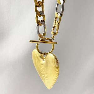 Op de afbeelding: Een goudkleurige ketting met een hartvormige hanger. De ketting is een mix van gouden en zilveren schakels. De hartvormige hanger heeft een gestructureerd, golvend ontwerp. De ketting heeft een tuimelsluiting. De sieraden worden tegen een witte achtergrond gepresenteerd.
