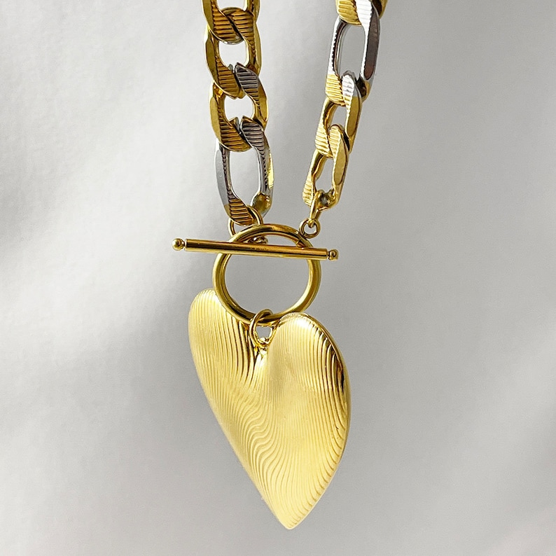 Op de afbeelding: Een goudkleurige ketting met een hartvormige hanger. De ketting is een mix van gouden en zilveren schakels. De hartvormige hanger heeft een gestructureerd, golvend ontwerp. De ketting heeft een tuimelsluiting. De sieraden worden tegen een witte achtergrond gepresenteerd.
