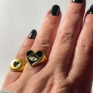 Pu&ograve; includere: Anelli dorati con dettagli in smalto nero. Un anello presenta una forma a cuore con un disegno a occhio, e l'altro ha un ritaglio a forma di cuore. Gli anelli sono mostrati su una mano con smalto nero.
