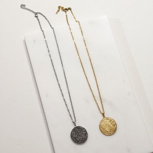Peut inclure: Deux colliers avec des pendentifs circulaires. L'un est argent&eacute;, l'autre dor&eacute;. Les deux ont une cha&icirc;ne d&eacute;licate et un pendentif au design complexe. Les colliers sont pr&eacute;sent&eacute;s sur une surface blanche.