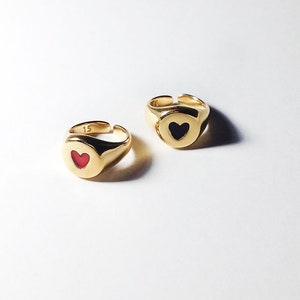 Pu&ograve; includere: Due anelli a sigillo color oro con disegni a forma di cuore. Un anello presenta un cuore rosso, l'altro un cuore nero. Gli anelli hanno una finitura lucida e sono su sfondo bianco. Il numero "15" &egrave; visibile su un anello.