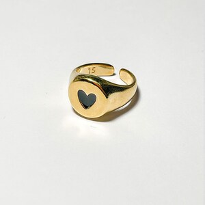 Pu&ograve; includere: Un anello regolabile color oro con un intarsio a forma di cuore nero. L'anello ha una finitura liscia e lucida e presenta il numero "15" inciso sulla fascia. L'anello &egrave; posto su uno sfondo bianco.