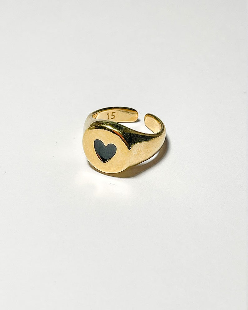 Pu&ograve; includere: Un anello regolabile color oro con un intarsio a forma di cuore nero. L'anello ha una finitura liscia e lucida e presenta il numero "15" inciso sulla fascia. L'anello &egrave; posto su uno sfondo bianco.