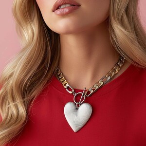 Op de afbeelding: Zilveren hartvormige hangerketting met een toggle sluiting. De ketting heeft een grove schakelketting met zilveren en goudkleurige schakels. De hartvormige hanger heeft een getextureerd, golvend ontwerp. De persoon draagt een rode top.