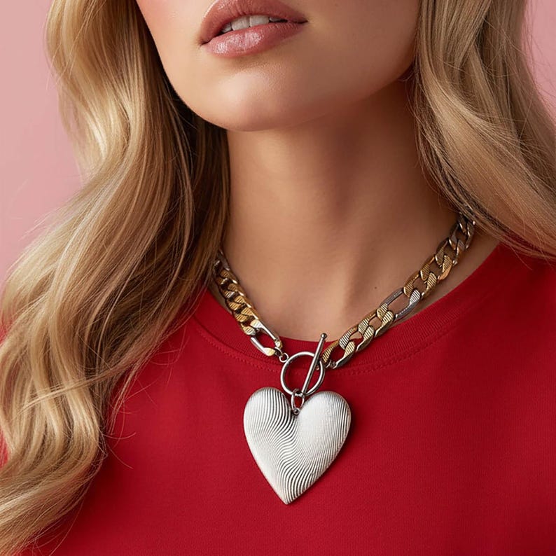Op de afbeelding: Zilveren hartvormige hangerketting met een toggle sluiting. De ketting heeft een grove schakelketting met zilveren en goudkleurige schakels. De hartvormige hanger heeft een getextureerd, golvend ontwerp. De persoon draagt een rode top.