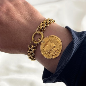 Può includere: Un braccialetto a catena dorata con un grande ciondolo a forma di moneta d'oro con un design dettagliato. La moneta è attaccata alla catena con un anello d'oro.