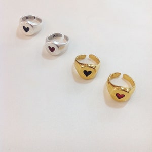 Pu&ograve; includere: Quattro anelli regolabili con facce a forma di cuore. Due sono argentati con un cuore nero, e due sono dorati con un cuore nero. Un anello d'argento ha inciso il numero "15". Gli anelli sono su una superficie bianca.