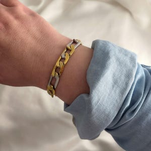 Può includere: Un braccialetto a catena bicolore oro e argento. Il braccialetto è composto da maglie interconnesse, con segmenti oro e argento alternati. È indossato su un polso, con una manica di camicia di jeans blu sullo sfondo.