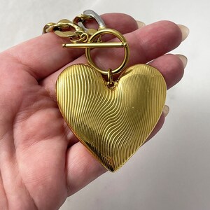 Op de afbeelding: Een goudkleurige hartvormige hanger met een gestructureerd, golvend ontwerp. De hanger is bevestigd aan een gouden ketting met een tuimelsluiting. De ketting heeft ook zilverkleurige schakels.