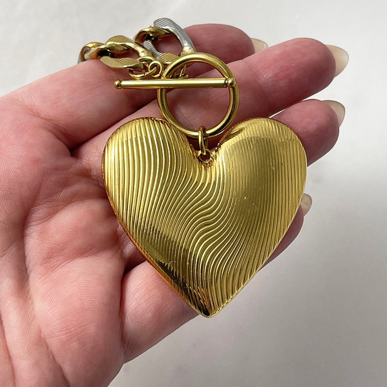 Op de afbeelding: Een goudkleurige hartvormige hanger met een gestructureerd, golvend ontwerp. De hanger is bevestigd aan een gouden ketting met een tuimelsluiting. De ketting heeft ook zilverkleurige schakels.