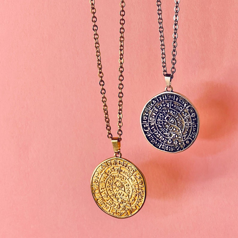 Peut inclure: Deux colliers pendentifs avec des pendentifs circulaires et des cha&icirc;nes. Un pendentif est dor&eacute;, l'autre est argent&eacute;. Les deux pendentifs pr&eacute;sentent un motif grav&eacute; avec une &eacute;criture ancienne. Le fond est rose uni.