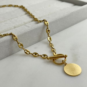 May include: A gold necklace with a toggle clasp and a circular pendant. The pendant has a sunburst design. The chain is made of oval-shaped links. The necklace is gold-coloured.
