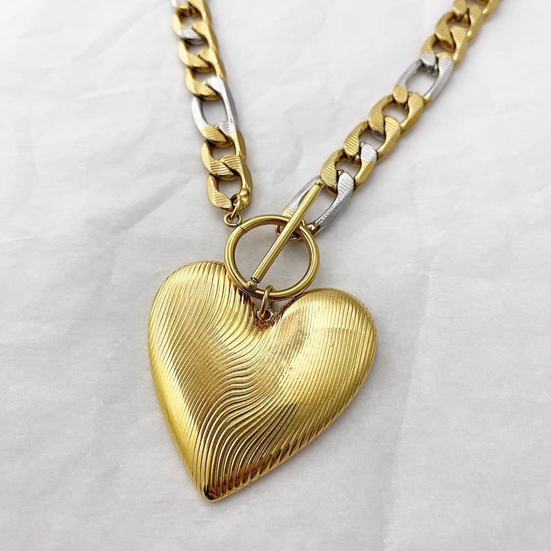 Op de afbeelding: Een goudkleurige hartvormige hangerketting met een getextureerd oppervlak. De ketting heeft een dikke ketting met afwisselend gouden en zilveren schakels. De tuimelsluiting is goudkleurig en het hart hangt aan een kleine ring.