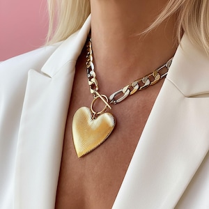 Op de afbeelding: Een goudkleurige hartvormige hangerketting met een getextureerd oppervlak. De ketting heeft een dikke ketting in goud- en zilverkleuren, vastgemaakt met een tuimelsluiting. De ketting wordt gedragen over een witte blazer.