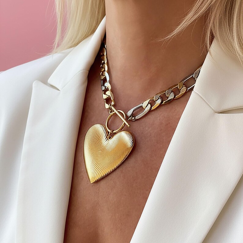Op de afbeelding: Een goudkleurige hartvormige hangerketting met een getextureerd oppervlak. De ketting heeft een dikke ketting in goud- en zilverkleuren, vastgemaakt met een tuimelsluiting. De ketting wordt gedragen over een witte blazer.