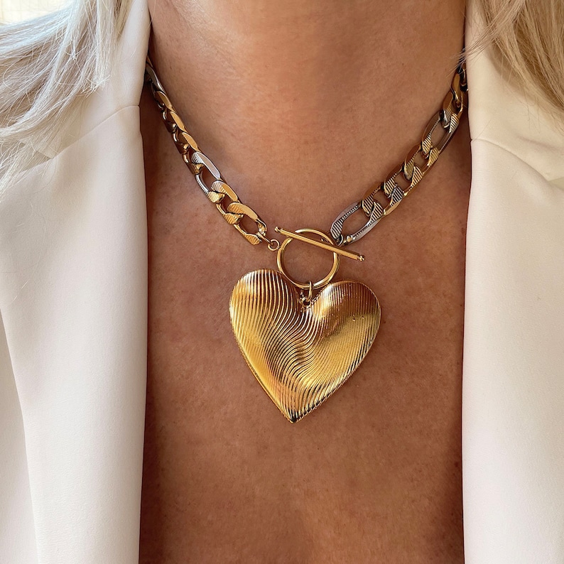 Op de afbeelding: Een goudkleurige ketting met een grote hartvormige hanger. Het hart heeft een gestructureerd, golvend ontwerp. De ketting is een mix van gouden en zilveren schakels, met een tuimelsluiting. De ketting wordt gedragen over een witte blazer.