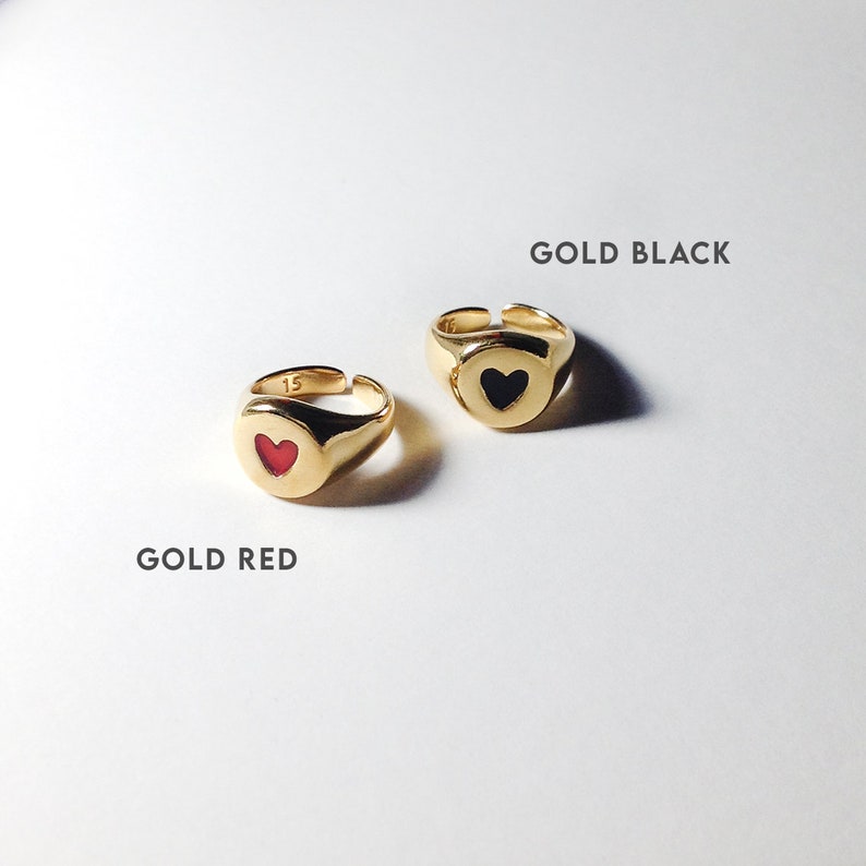 Pu&ograve; includere: Due anelli a sigillo dorati con cuori al centro. Un anello ha un cuore rosso, l'altro un cuore nero. Gli anelli sono esposti su una superficie bianca, con i testi "GOLD RED" e "GOLD BLACK".