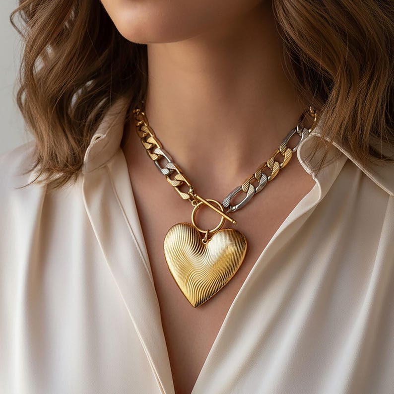 Op de afbeelding: Een goudkleurige hartvormige hangerketting met een getextureerd oppervlak. De ketting heeft een grove ketting met gouden en zilveren schakels en een tuimelsluiting. De hartvormige hanger is groot en prominent, waardoor een opvallend sieraad ontstaat.