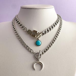 Op de afbeelding: Set van zilveren kettingkettingen met twee lagen. De bovenste laag heeft een slot met een turquoise stenen hanger. De onderste laag heeft een halve maan hanger.