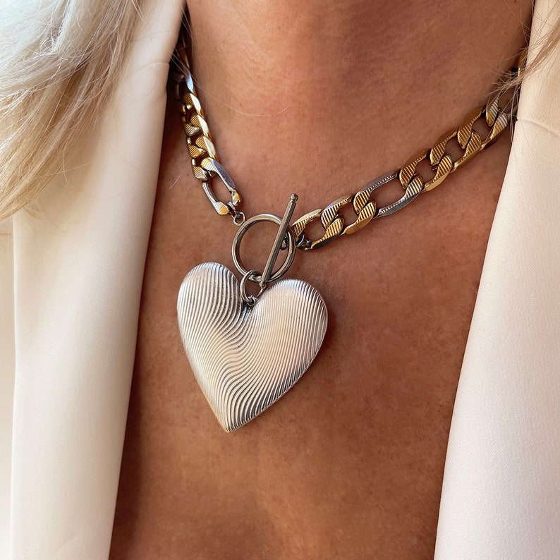 Op de afbeelding: Een zilveren hartvormige hangerketting met een getextureerd oppervlak. De ketting heeft een dikke ketting met zilveren en gouden schakels en een tuimelsluiting. De hartvormige hanger is het middelpunt van de ketting.