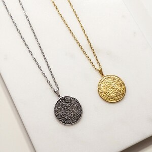 Peut inclure: Deux colliers avec des pendentifs circulaires. L'un est argent&eacute;, l'autre dor&eacute;. Chaque pendentif pr&eacute;sente un motif grav&eacute; et est suspendu &agrave; une fine cha&icirc;ne. Les colliers sont pr&eacute;sent&eacute;s sur une surface en marbre blanc.