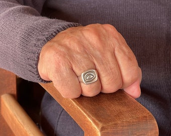 Può includere: Un anello d'argento con una faccia quadrata con un disegno a occhio. L'anello è indossato su una mano appoggiata sul bracciolo di una sedia di legno. La persona indossa un maglione grigio.