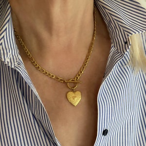 Peut inclure: Un collier doré avec un pendentif en forme de cœur. Le pendentif présente un motif orné et est suspendu à une chaîne avec un fermoir à bascule. Le collier est porté sur une chemise rayée bleue et blanche.