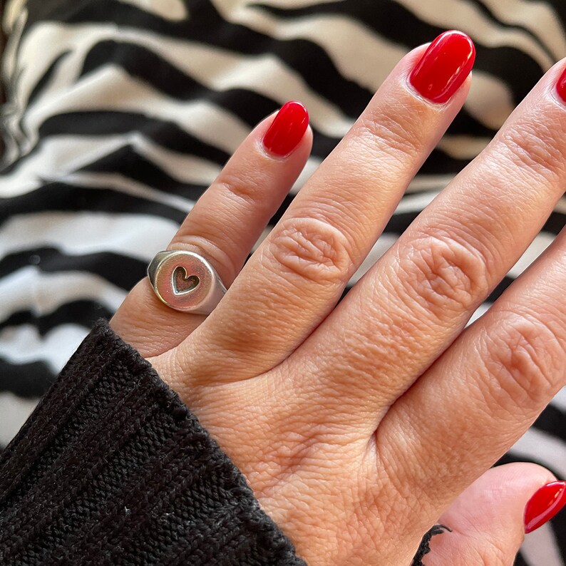 Pu&ograve; includere: Un anello d'argento con un cuore traforato. L'anello &egrave; indossato su un dito con smalto rosso. Lo sfondo &egrave; una stampa zebrata in bianco e nero.