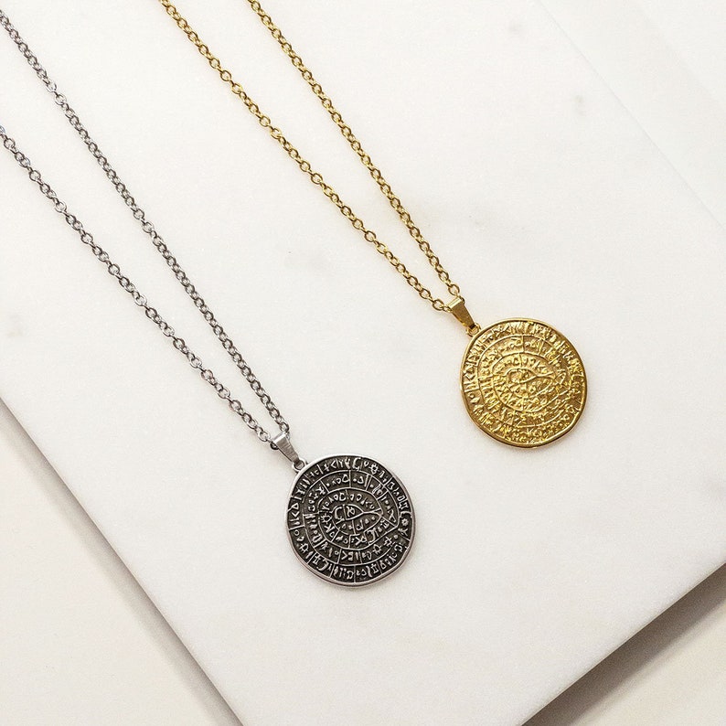 Peut inclure: Deux colliers avec des pendentifs circulaires. L'un est argent&eacute;, l'autre dor&eacute;. Les deux ont des cha&icirc;nes d&eacute;licates. Les pendentifs pr&eacute;sentent un motif grav&eacute;. Les colliers sont pr&eacute;sent&eacute;s sur une surface en marbre blanc.