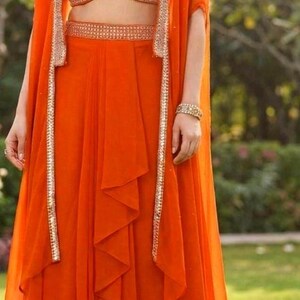May include: A vibrant orange three-piece ensemble featuring a sleeveless cropped bodice with white bird embroidery, a matching high-waisted skirt with a ruffled front panel, and a long, open-front cape with metallic silver trim along the edges.