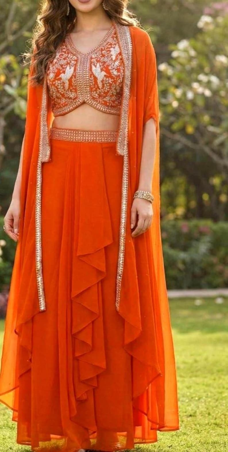 May include: A vibrant orange three-piece ensemble featuring a sleeveless cropped bodice with white bird embroidery, a matching high-waisted skirt with a ruffled front panel, and a long, open-front cape with metallic silver trim along the edges.