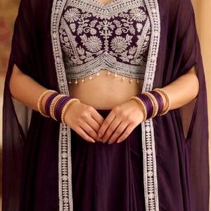 May include: A deep purple ensemble featuring a cropped bodice with intricate silver floral embroidery and a matching sheer cape. The outfit is accessorized with a gold-toned beaded necklace and multiple purple bangles on each wrist.