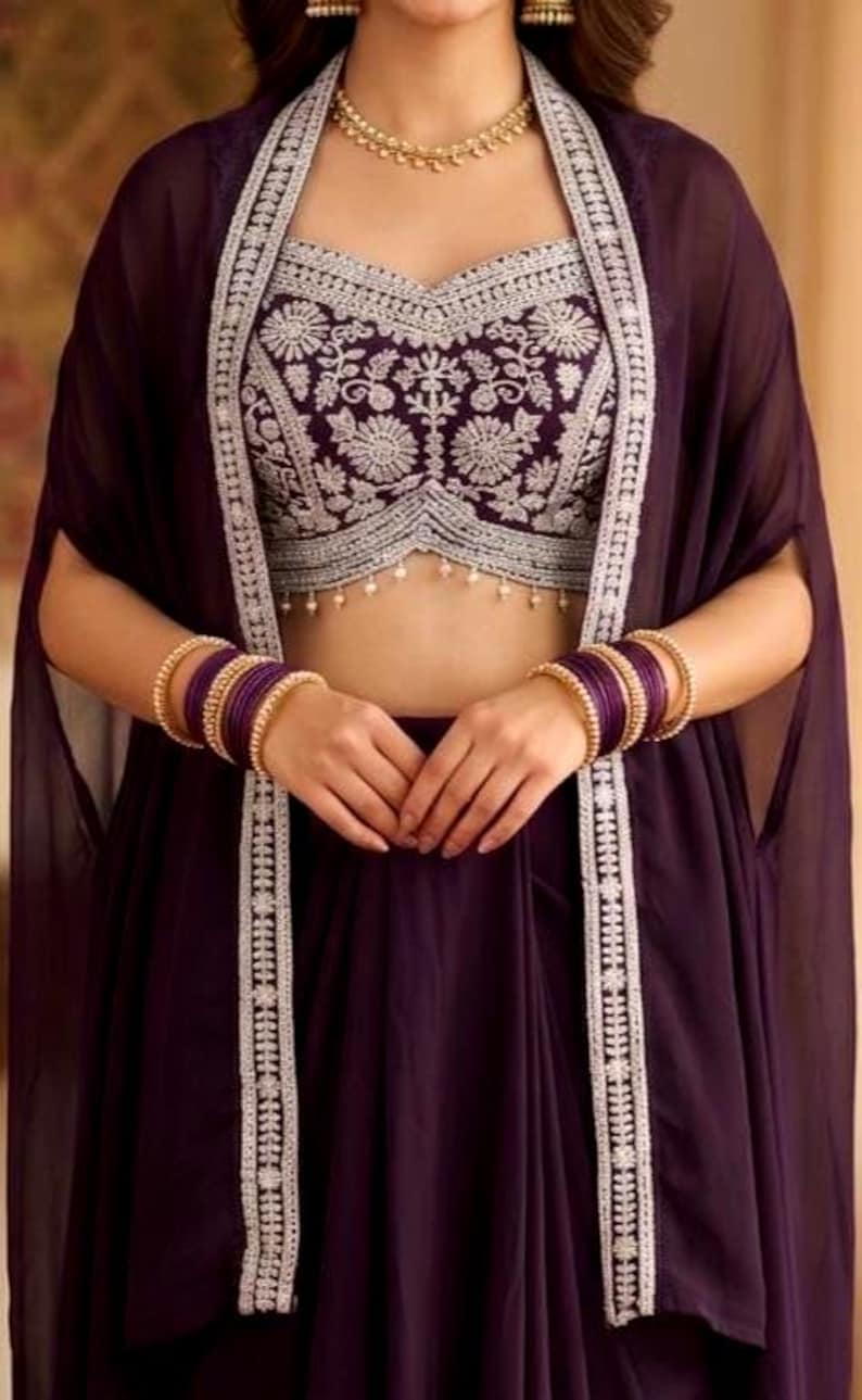 May include: A deep purple ensemble featuring a cropped bodice with intricate silver floral embroidery and a matching sheer cape. The outfit is accessorized with a gold-toned beaded necklace and multiple purple bangles on each wrist.