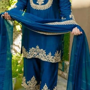 May include: A deep teal blue three-piece outfit consisting of a long-sleeved tunic, wide-leg trousers, and a matching sheer dupatta. The tunic and trousers feature intricate gold embroidery along the neckline, cuffs, and hemline.