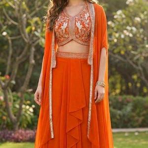 May include: A vibrant orange three-piece ensemble consisting of a sleeveless cropped top, a high-waisted skirt, and a long open-front cape. The top features intricate white embroidery and silver embellishments, while the cape and skirt have matching silver trim along the edges.