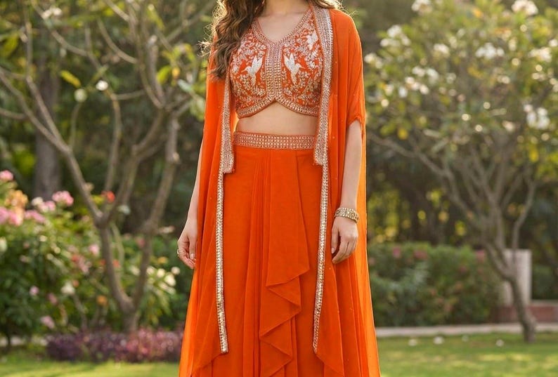May include: A vibrant orange three-piece ensemble consisting of a sleeveless cropped top, a high-waisted skirt, and a long open-front cape. The top features intricate white embroidery and silver embellishments, while the cape and skirt have matching silver trim along the edges.