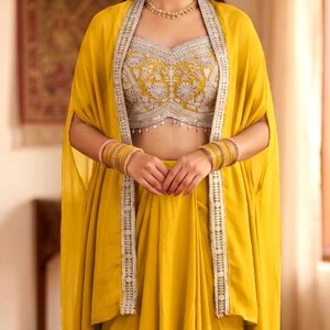 May include: A mustard yellow ensemble featuring a sleeveless crop top with intricate silver embroidery and hanging beads, paired with a matching floor-length skirt and a long, open-front cape with silver-trimmed edges and gold bangles.