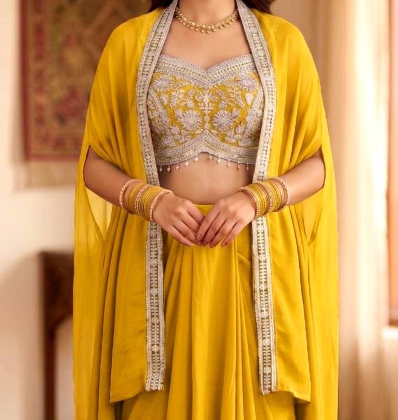 May include: A mustard yellow ensemble featuring a sleeveless crop top with intricate silver embroidery and hanging beads, paired with a matching floor-length skirt and a long, open-front cape with silver-trimmed edges and gold bangles.