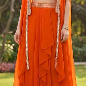 May include: A vibrant orange three-piece ensemble consisting of a sleeveless cropped bodice with white bird embroidery, a matching high-waisted skirt with vertical ruffled detailing, and a long, open-front cape featuring metallic silver trim.
