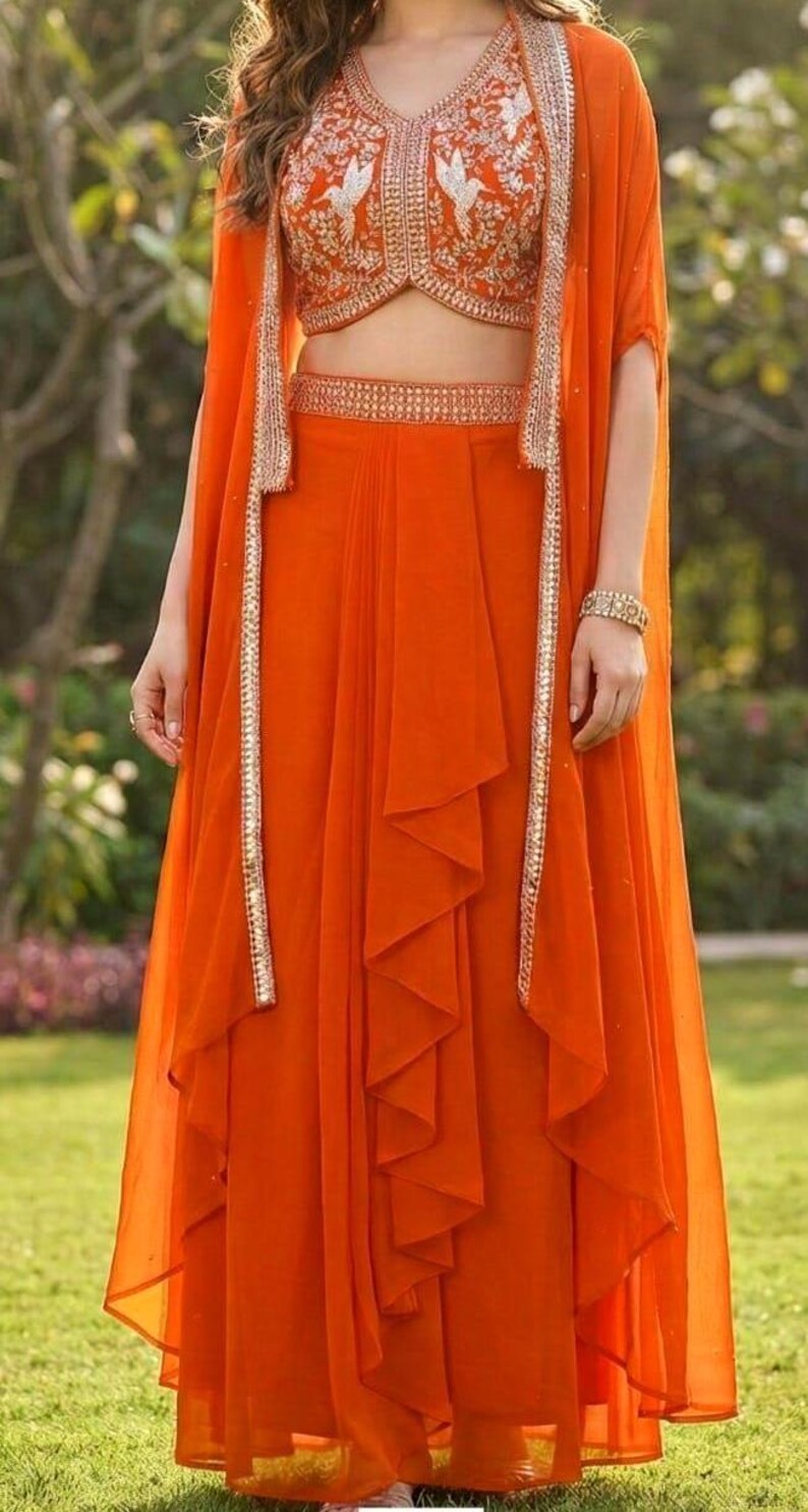 May include: A vibrant orange three-piece ensemble consisting of a sleeveless cropped bodice with white bird embroidery, a matching high-waisted skirt with vertical ruffled detailing, and a long, open-front cape featuring metallic silver trim.