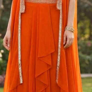 May include: A vibrant orange three-piece ensemble featuring a sleeveless cropped bodice with white bird embroidery, a matching high-waisted floor-length skirt with a central ruffled panel, and a long, open-front cape with metallic silver trim.