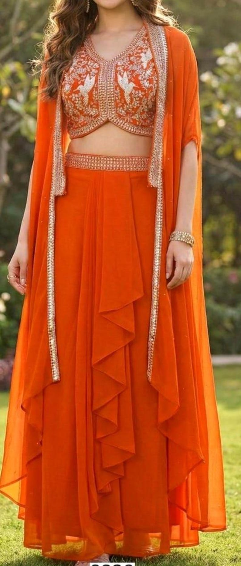 May include: A vibrant orange three-piece ensemble featuring a sleeveless cropped bodice with white bird embroidery, a matching high-waisted floor-length skirt with a central ruffled panel, and a long, open-front cape with metallic silver trim.