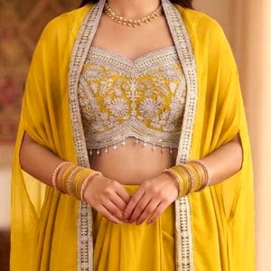 May include: A bright yellow sleeveless crop top featuring intricate silver embroidery and a scalloped hem with small hanging beads, paired with a matching yellow cape-style overlay and stacked gold bangles on both wrists.