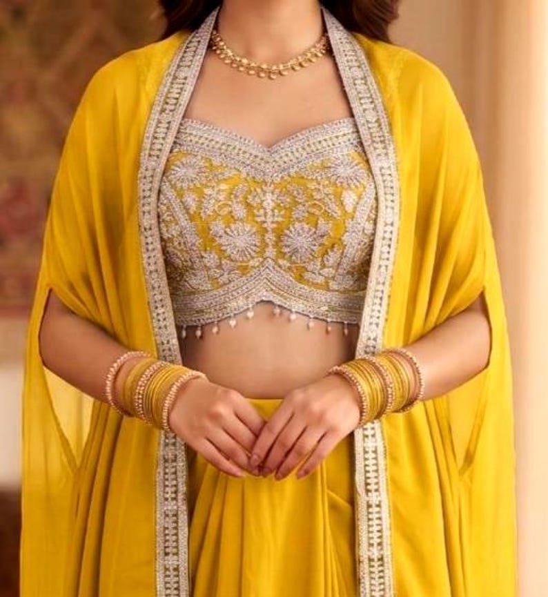 May include: A bright yellow sleeveless crop top featuring intricate silver embroidery and a scalloped hem with small hanging beads, paired with a matching yellow cape-style overlay and stacked gold bangles on both wrists.