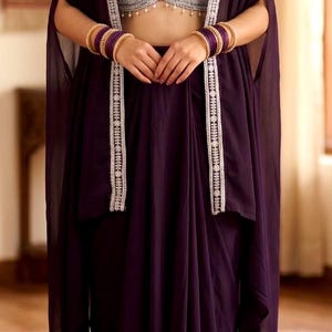 May include: A deep purple ensemble consisting of a sleeveless crop top with intricate silver floral embroidery and a matching long skirt. A sheer purple cape with silver embroidered trim is worn over the top, paired with gold bangles and a necklace.