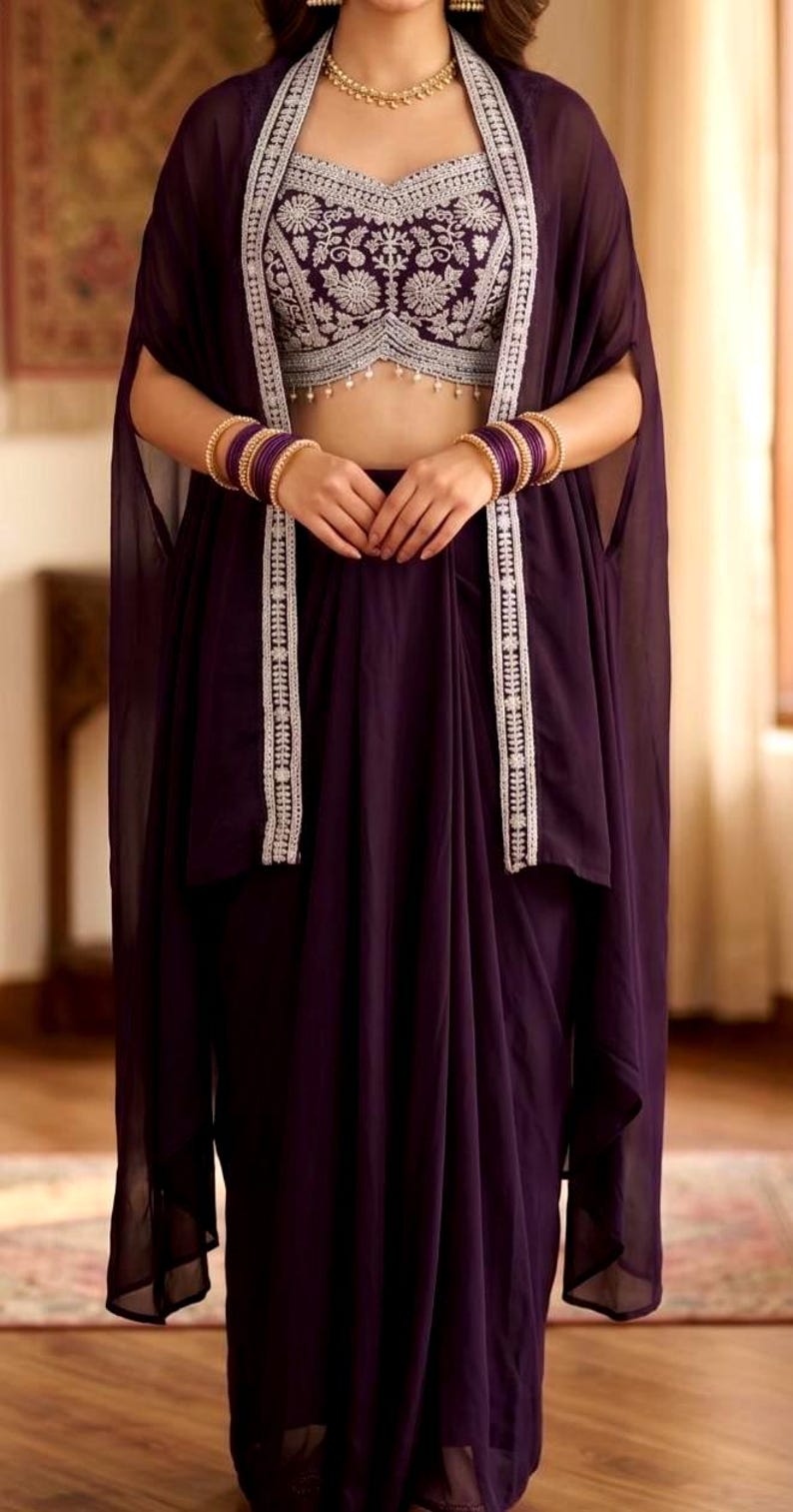 May include: A deep purple ensemble consisting of a sleeveless crop top with intricate silver floral embroidery and a matching long skirt. A sheer purple cape with silver embroidered trim is worn over the top, paired with gold bangles and a necklace.
