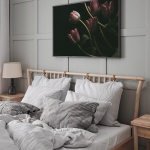 May include: A bedroom with a wooden bed frame and white bedding. The headboard has leather straps. There are several pillows on the bed, including a striped pillow. A framed print of pink tulips on a dark background hangs on the wall above the bed.