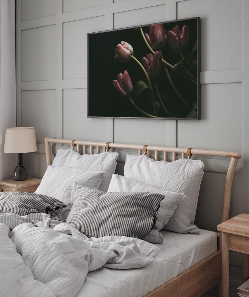 May include: A bedroom with a wooden bed frame and white bedding. The headboard has leather straps. There are several pillows on the bed, including a striped pillow. A framed print of pink tulips on a dark background hangs on the wall above the bed.