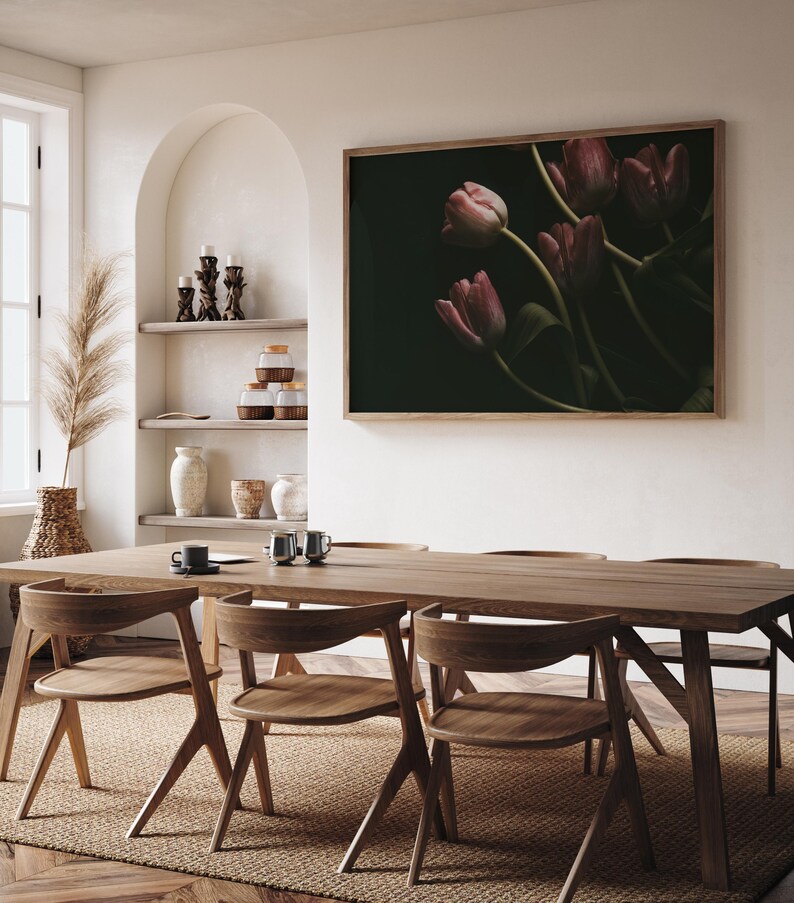 May include: A wooden dining table with four chairs, set against a white wall with a framed print of pink tulips on a dark background. The table is made of natural wood with a light brown finish. The chairs are also made of wood with a similar finish. The print is framed in a dark wood frame.